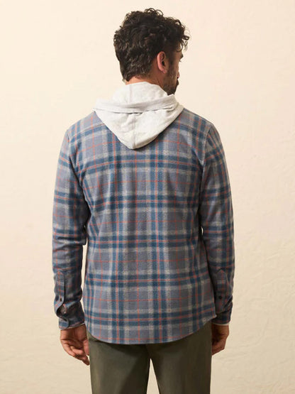 A man faces away from the camera, wearing a gray hoodie under a Faherty Brand Legend Sweater Shirt in Three Lakes Plaid and green pants, set against a plain beige background.