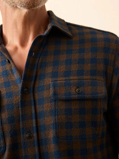 Close-up of a person wearing the Faherty Brand Legend Sweater Shirt in Woodland Check, showing its blue and brown checkered pattern, front pocket, and soft knit texture, with part of their neck and chin visible.