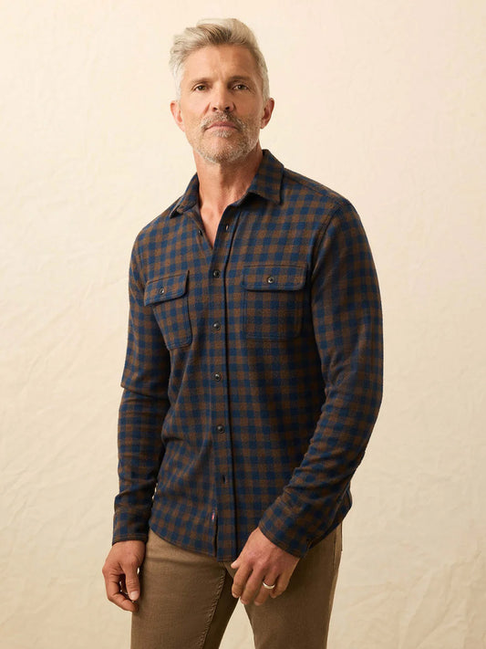 A man with short gray hair and a beard wears a Faherty Brand Legend Sweater Shirt in Woodland Check and tan pants, standing against a plain beige background.