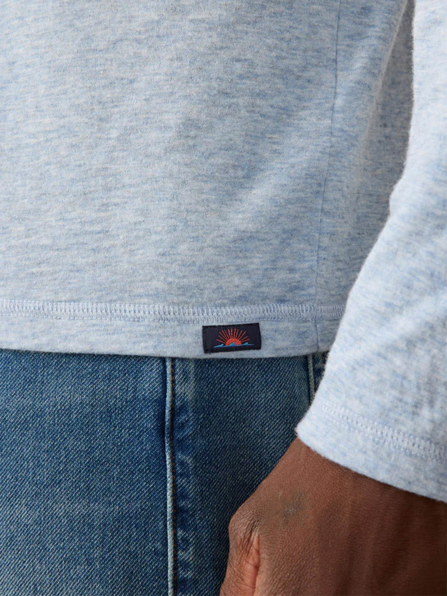 Close-up of a person wearing the Faherty Brand Cloud Cotton Long-Sleeve Henley in Light Blue Heather with blue jeans, highlighting a small fabric tag with a red sunburst logo near the shirt’s hem.
