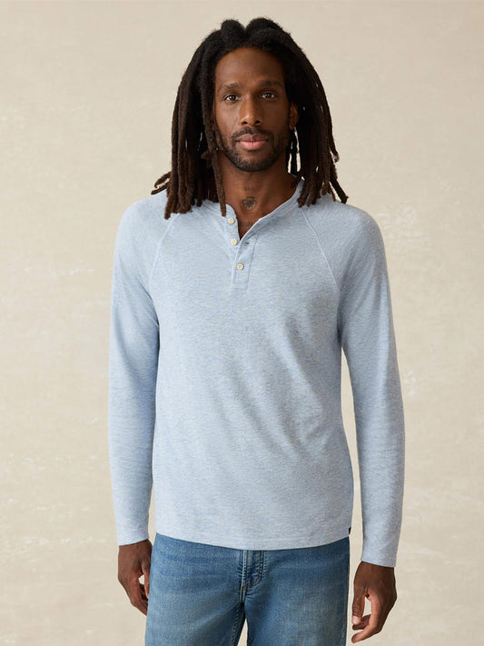 A man with long dreadlocks is wearing the Faherty Brand Cloud Cotton Long-Sleeve Henley in Light Blue Heather and blue jeans, standing against a plain beige background.