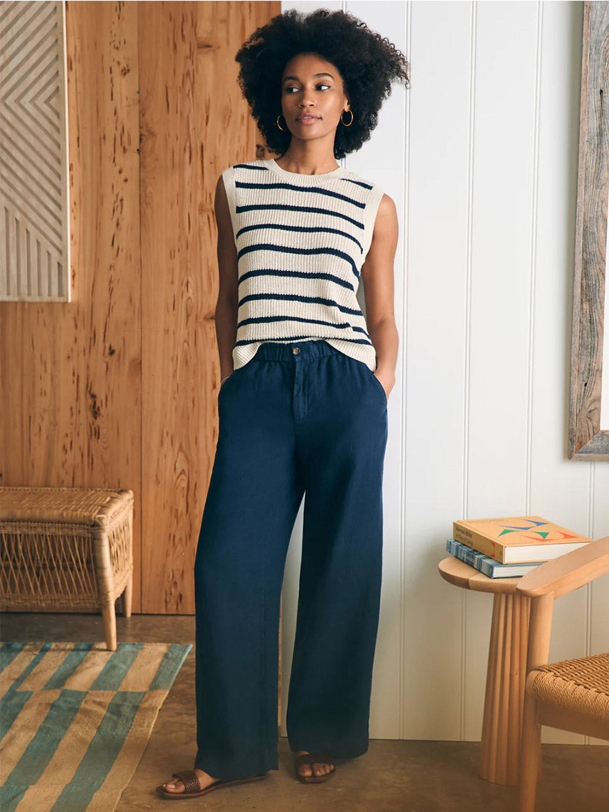 A woman in a striped sleeveless top and Faherty Brand Monterey Linen Pant in After Midnight stands in a wooden paneled room, hands in pockets.