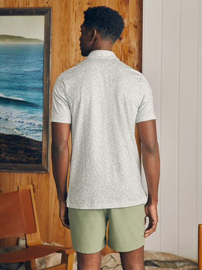 Man in a Faherty Brand Movement Short-Sleeve Pique Polo Sky Canopy Print with vented hems and green shorts standing in front of a seaside painting.