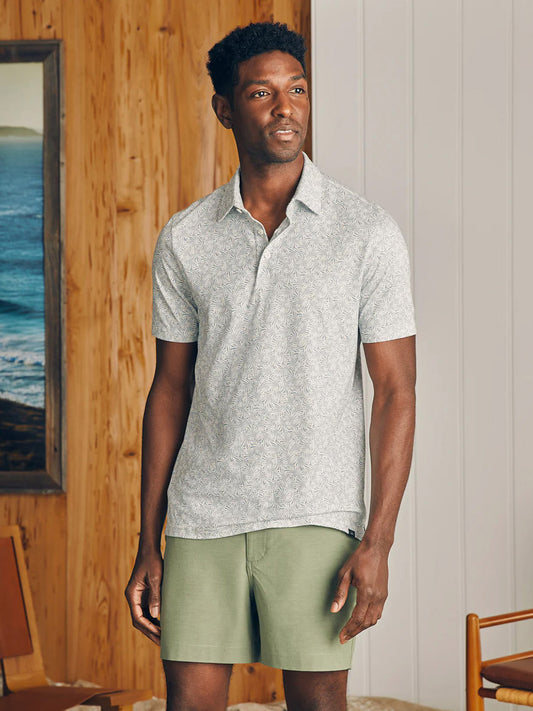 A man in a Faherty Brand Movement Short-Sleeve Pique Polo Sky Canopy Print crafted from innovative fabrics and green shorts standing indoors with a coastal view through the window behind him.