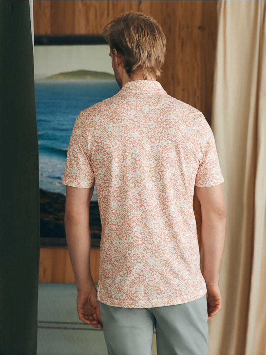 Man standing indoors looking towards a window with a view of the sea, wearing a Faherty Brand Movement Short-Sleeve Polo in Hilo Rose Floral Print made of innovative fabrics and light trousers with vented hems.