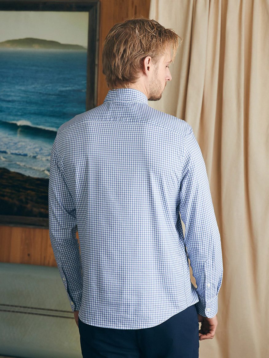 A person with blonde hair, seen from behind, wearing a Faherty Brand Movement Shirt in Light Blue Gingham made of wrinkle-resistant Supima cotton, stands indoors near a beige curtain and a framed seascape painting.