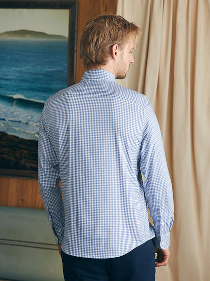 A person with blonde hair, seen from behind, wearing a Faherty Brand Movement Shirt in Light Blue Gingham made of wrinkle-resistant Supima cotton, stands indoors near a beige curtain and a framed seascape painting.