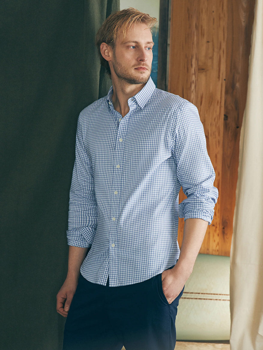 A man with blonde hair is standing indoors wearing a Faherty Brand Movement Shirt in light blue gingham and dark pants, with his sleeves rolled up. He is looking to his left.