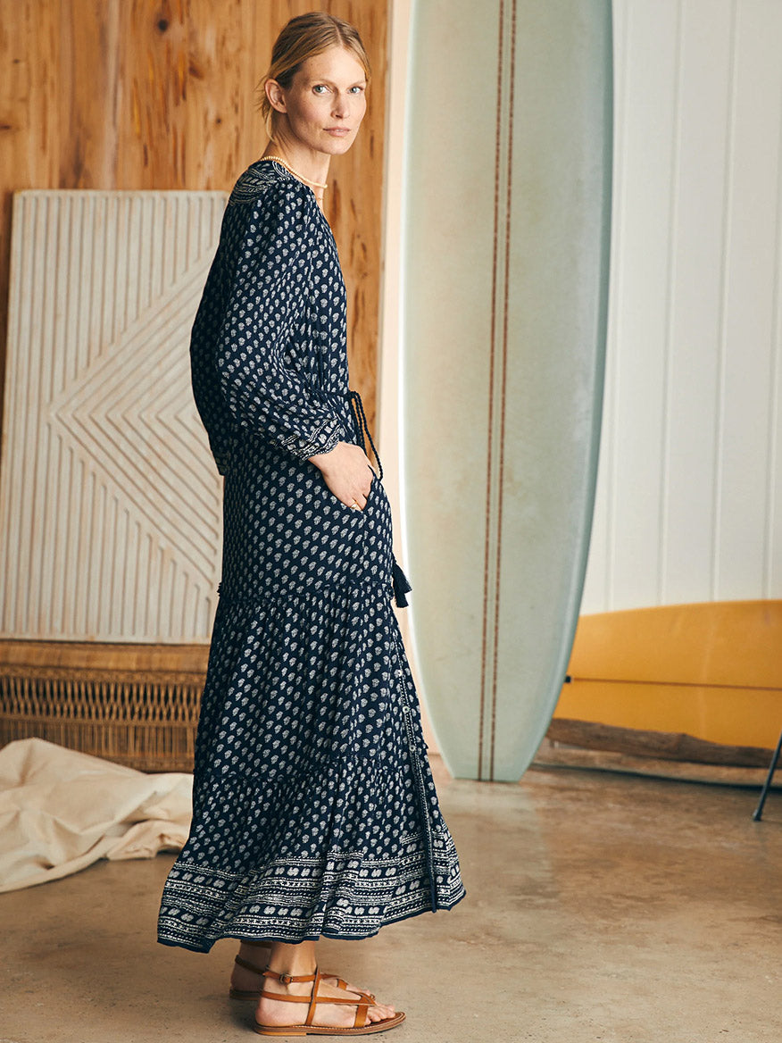 A woman in a Faherty Brand Orinda Long Sleeve Maxi Dress in Lotus Floral Print standing in a room with surfboards leaning against the wall.