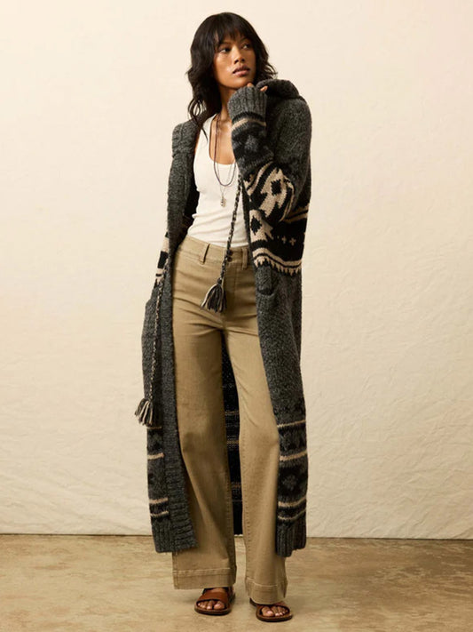 A woman stands indoors in the Faherty Brand Steven Paul Judd Chahtah Hooded Duster in Charcoal Chahtah, a white tank top, beige wide-leg pants, and sandals, with one hand touching her collar.