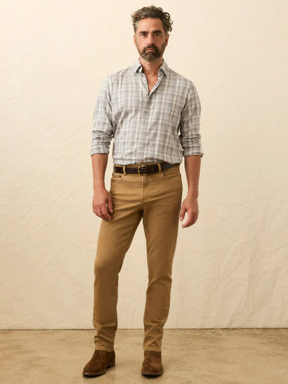 A man with short wavy hair and a beard wears a light gray plaid shirt, Faherty Brand Stretch Terry 5-Pocket Pant in Elk Brown, a brown belt, and brown shoes, standing against a neutral background.