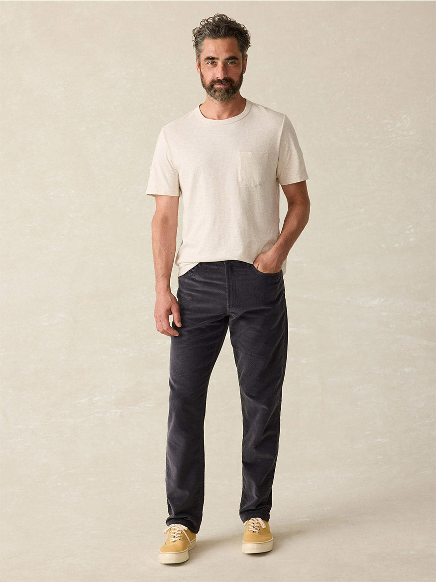 A man with short hair and a beard stands with one hand in his pocket, wearing a light t-shirt, Faherty Brand Stretch Terry Corduroy 5-Pocket Pant in Washed Charcoal, and yellow sneakers against a plain background.