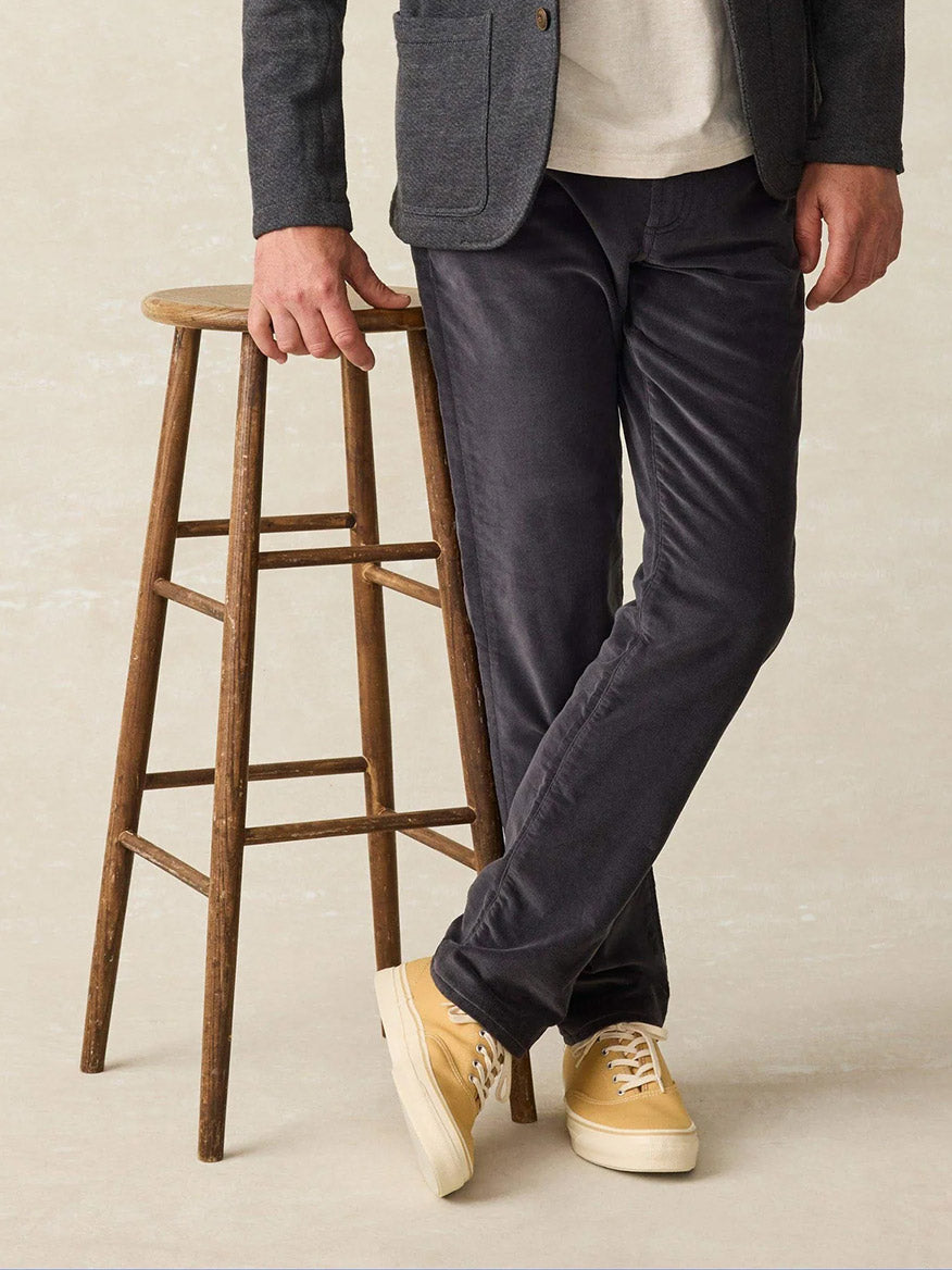 A person in a gray blazer and Faherty Brand Stretch Terry Corduroy 5-Pocket Pant in Washed Charcoal leans against a wooden stool, with yellow sneakers completing the classic fit.