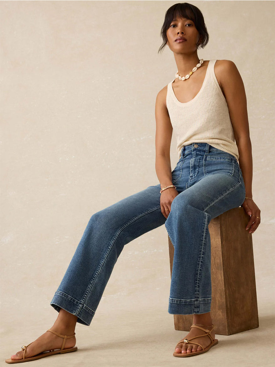 A woman sits on a wooden block wearing a beige slub knit tank top, Faherty Brand Stretch Terry 5-Pocket Ankle Pant in Riverton, tan sandals, and a pearl necklace.