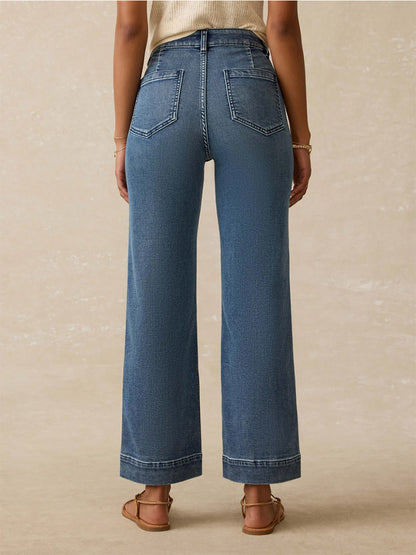 Wearing Faherty Brand Stretch Terry 5-Pocket Ankle Pant in Riverton, a person stands facing away from the camera in relaxed fit blue jeans and sandals against a neutral background.