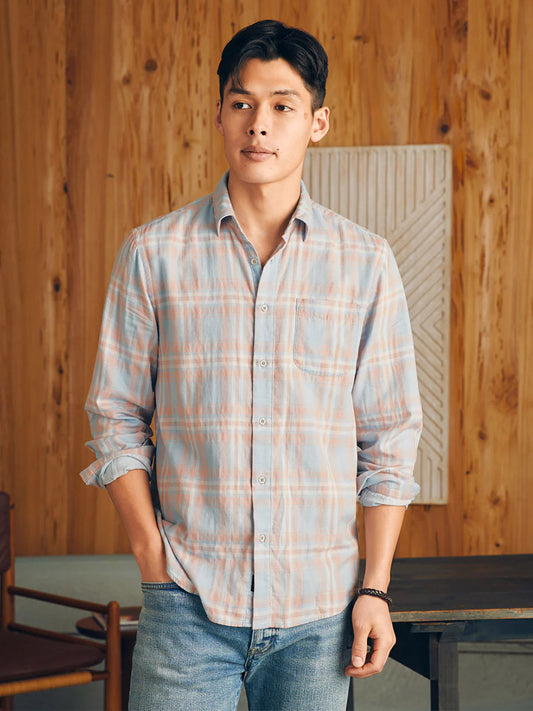 A man wearing a Faherty Brand Sunwashed Chambray Shirt in Coral Bay Plaid and jeans standing in a room with wooden paneling.