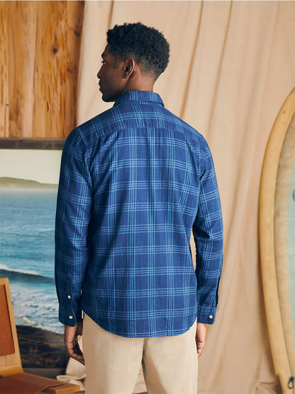 Man wearing a Faherty Brand Sunwashed Chambray Shirt in Navy Night Windowpane standing in a room with a surfboard and a painting of the sea in the background.