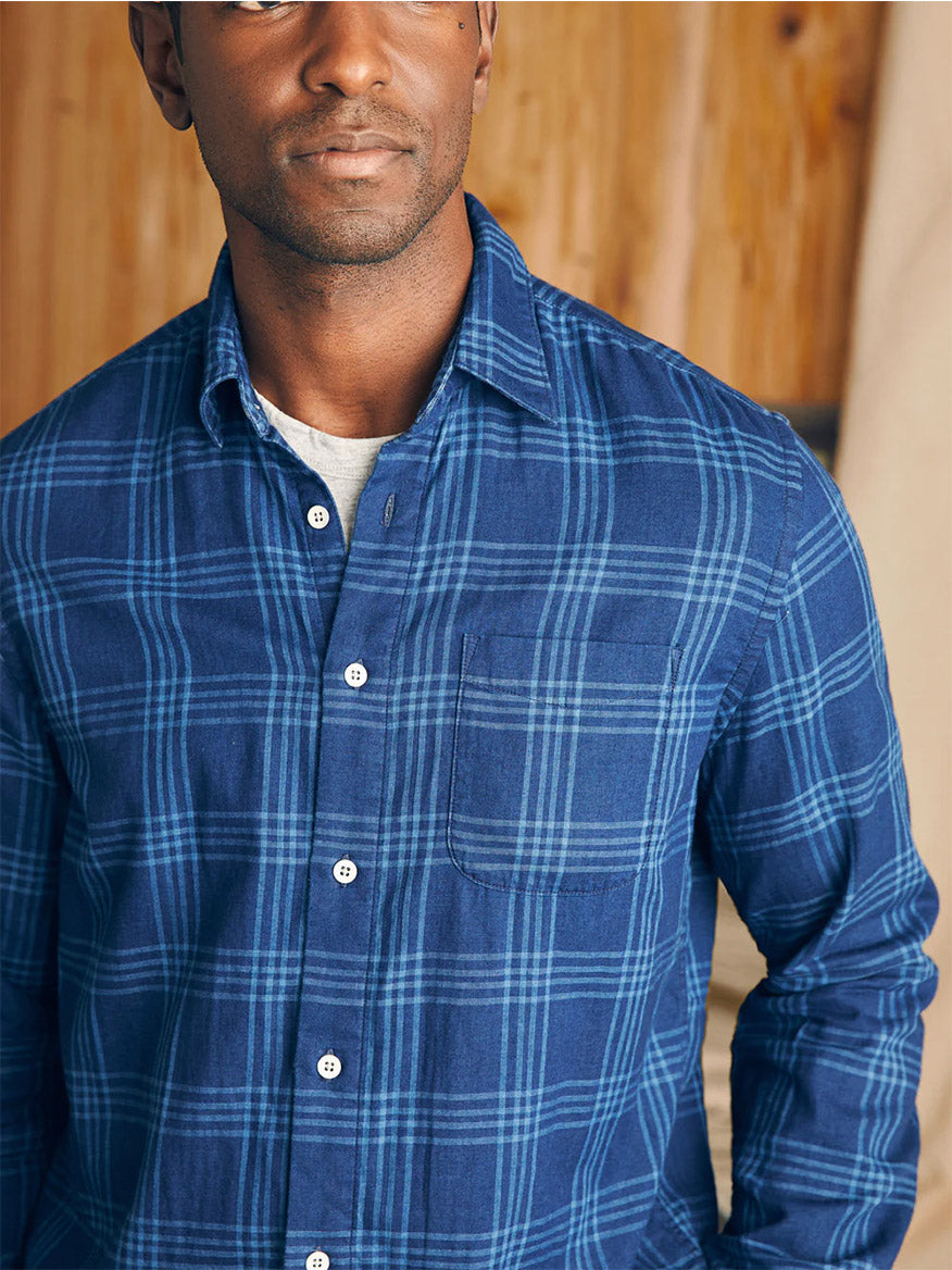 Man wearing a Faherty Brand Sunwashed Chambray Shirt in Navy Night Windowpane standing against a wooden background.