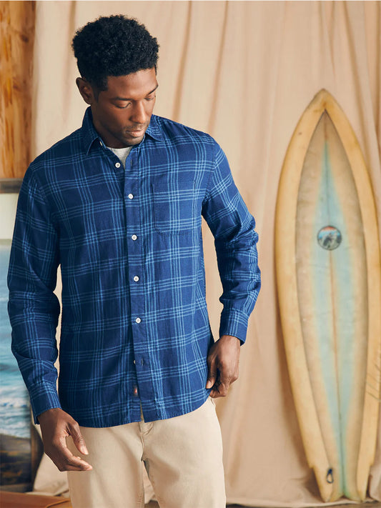 A man wearing a Faherty Brand Sunwashed Chambray Shirt in Navy Night Windowpane and beige pants is standing in a room with a surfboard in the background, ready for a casual evening.