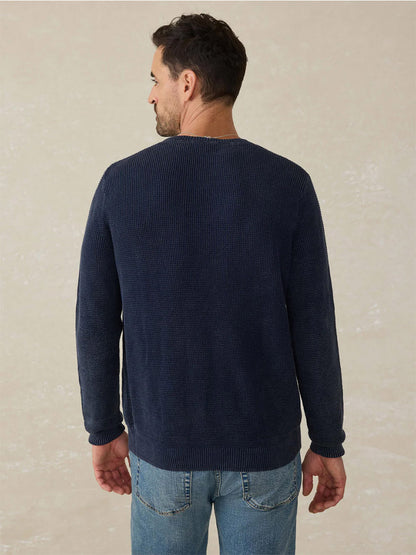 A man stands with his back to the camera, wearing the Faherty Brand Sunwashed Crewneck Sweater in Blue Nights and light blue jeans against a neutral background for a timeless, vintage look.