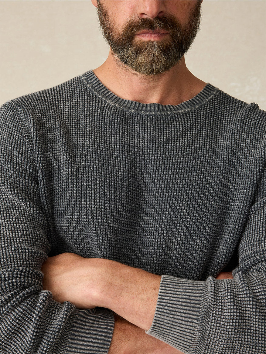 A bearded man wearing a Faherty Brand Sunwashed Crewneck Sweater in Faded Black stands with his arms crossed, exuding a vintage look.