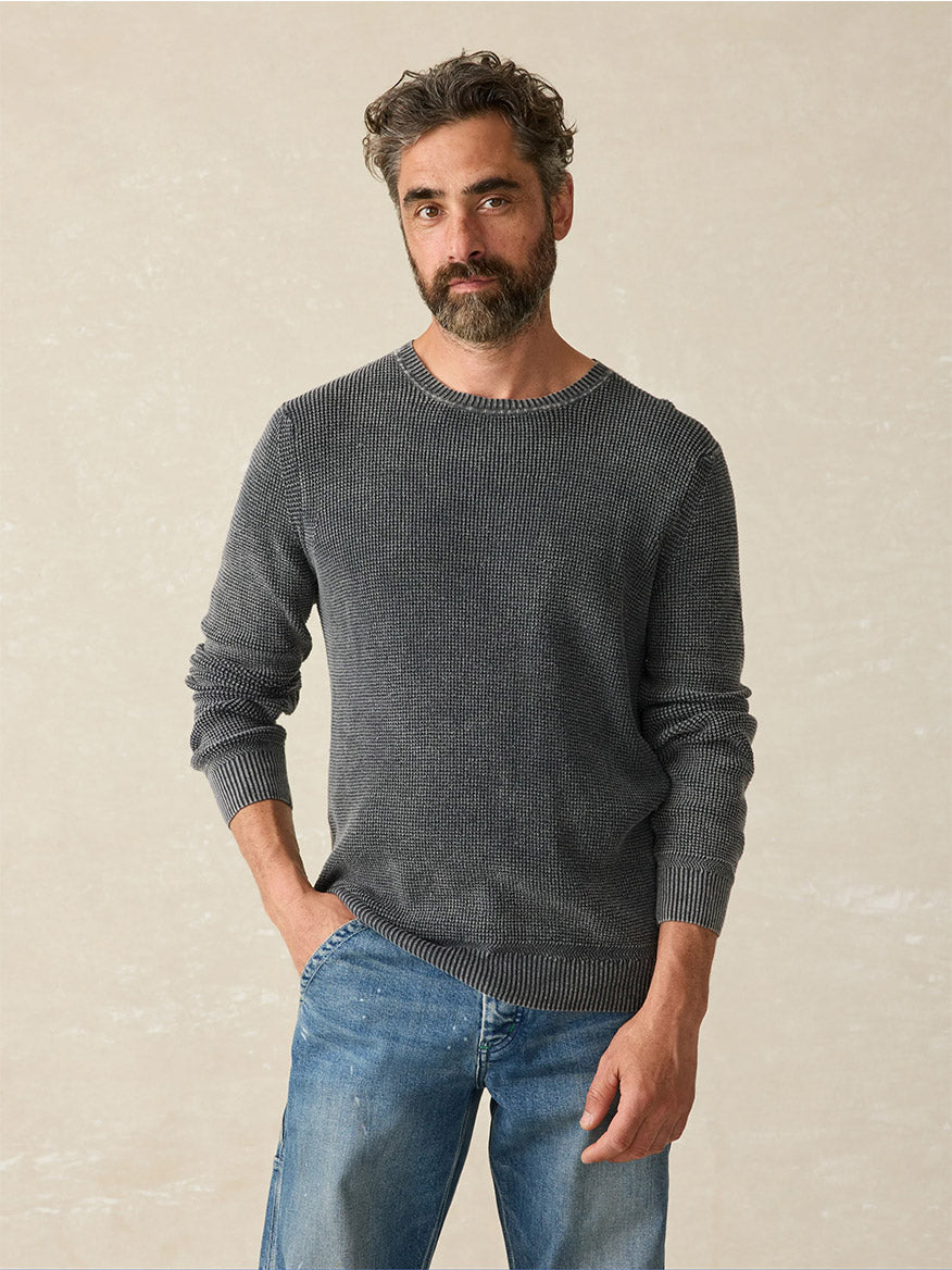 A man with a beard and curly hair wears the Faherty Brand Sunwashed Crewneck Sweater in Faded Black and blue jeans, posing against a textured beige background for a vintage look.