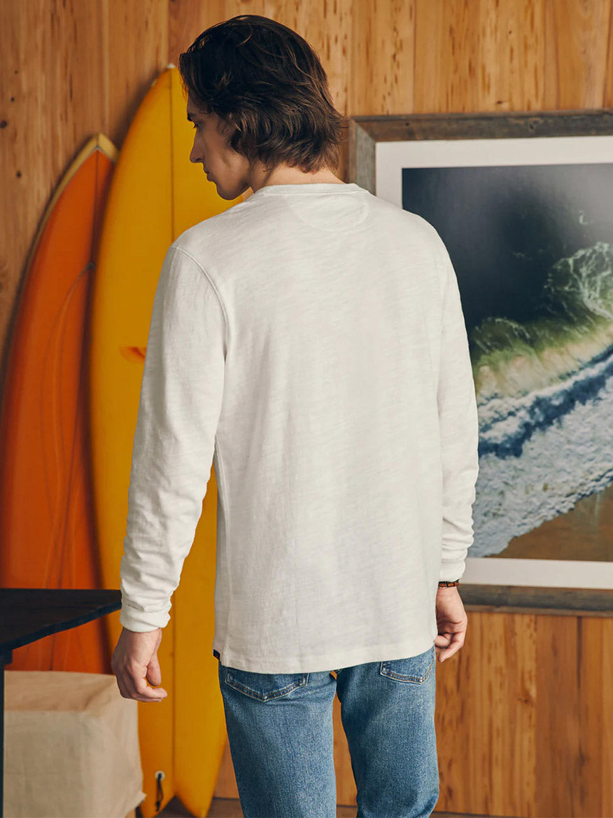 Man standing indoors, viewed from behind, wearing a Faherty Brand Sunwashed Slub Henley in White made of high-quality cotton and blue jeans, with surfboards and a framed wave photograph in the background.