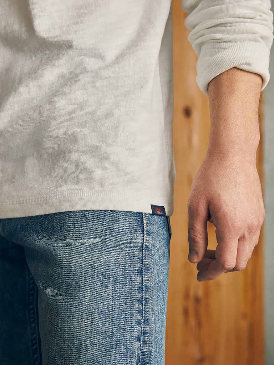 Man wearing a Faherty Brand Sunwashed Slub Henley in White and blue jeans standing against a wooden background.