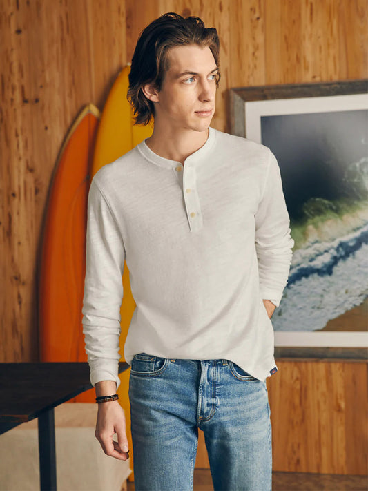 A man in a Faherty Brand Sunwashed Slub Henley in White and jeans standing indoors with a surfboard in the background.