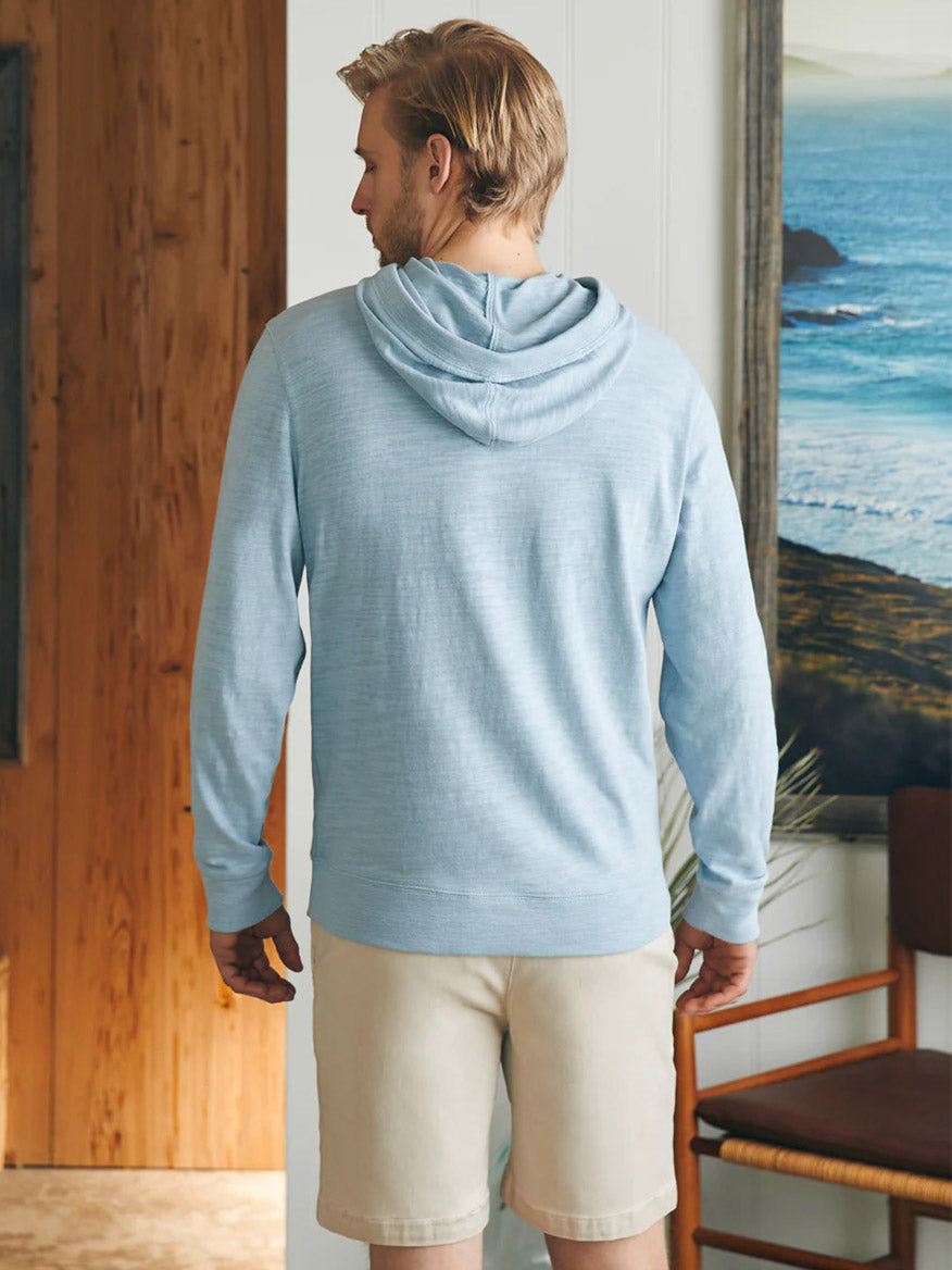 Man in a Faherty Brand Sunwashed Slub Hoodie in Blue Breeze and beige shorts standing by a window overlooking the ocean.