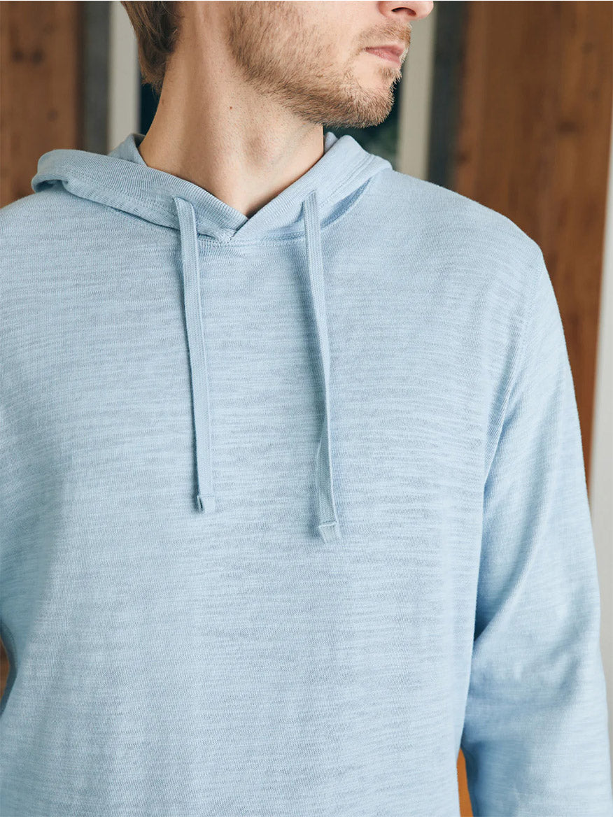 Man wearing a Faherty Brand Sunwashed Slub Hoodie in Blue Breeze made of American-grown cotton, featuring a blue hooded sweatshirt with drawstring detail.