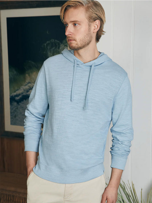 Man in a Faherty Brand Sunwashed Slub Hoodie in Blue Breeze, made from American-grown cotton, looking to the side with a painting in the background.