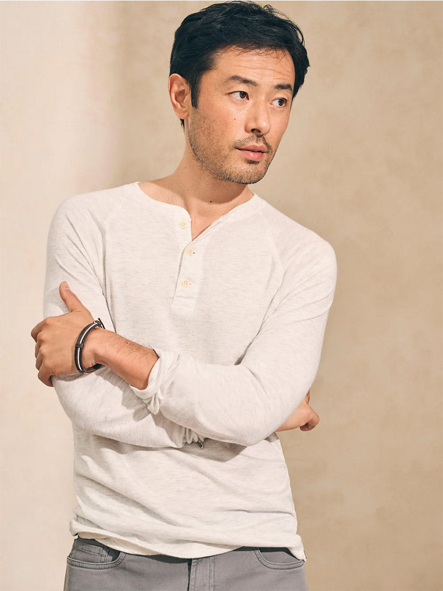 A man in a Faherty Brand Cloud Cotton Long-Sleeve Henley in Ivory Heather, with his arms crossed and gazing thoughtfully to the side against a neutral background.