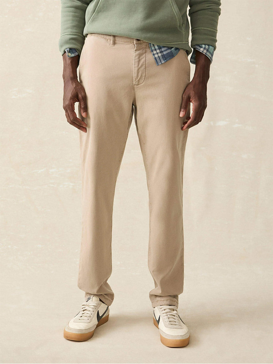 A person stands wearing versatile Faherty Brand Coastline Stretch Chino pants in Utility Khaki, a green hoodie, a blue plaid shirt, and white sneakers against a plain background.