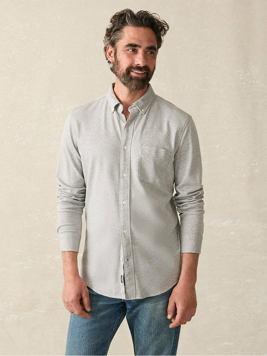 A bearded man in a grey heather twill Coastline Knit Shirt by Faherty Brand and blue jeans stands against a neutral background.