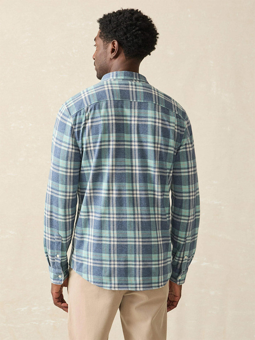 A person wearing the Faherty Brand Coastline Knit Shirt in Holbrook Island Plaid, renowned for its exceptional comfort, along with beige pants stands facing away against a neutral background. The wardrobe choice emphasizes wearability, showcasing a relaxed yet stylish appeal perfect for any setting.