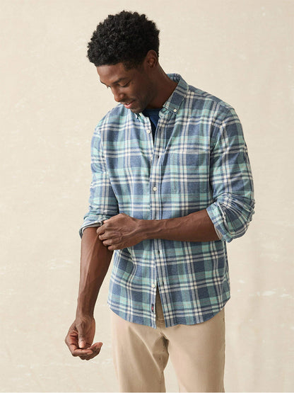 Dressed in the Faherty Brand Coastline Knit Shirt in Holbrook Island Plaid, a person emanates comfort and style as they roll up their sleeves, thoughtfully gazing down against a neutral backdrop.