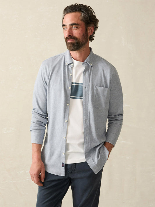 A man wearing a Faherty Brand Coastline Knit Shirt in Oceanside Blue over a white t-shirt with a central stripe, posing against a plain background.