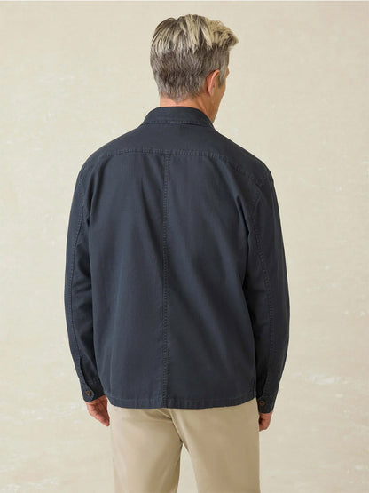 A man with short, light hair is seen from behind wearing the Faherty Brand Commander Shirt Jacket in Washed Charcoal, made from breathable organic cotton-linen for durability, along with beige pants, standing against a neutral background.