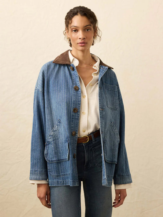 A woman faces forward, wearing the Faherty Brand Sunwashed Denim Barn Jacket in Dusk Stripe Wash with large front pockets and a corduroy collar, layered over a white shirt and dark jeans.