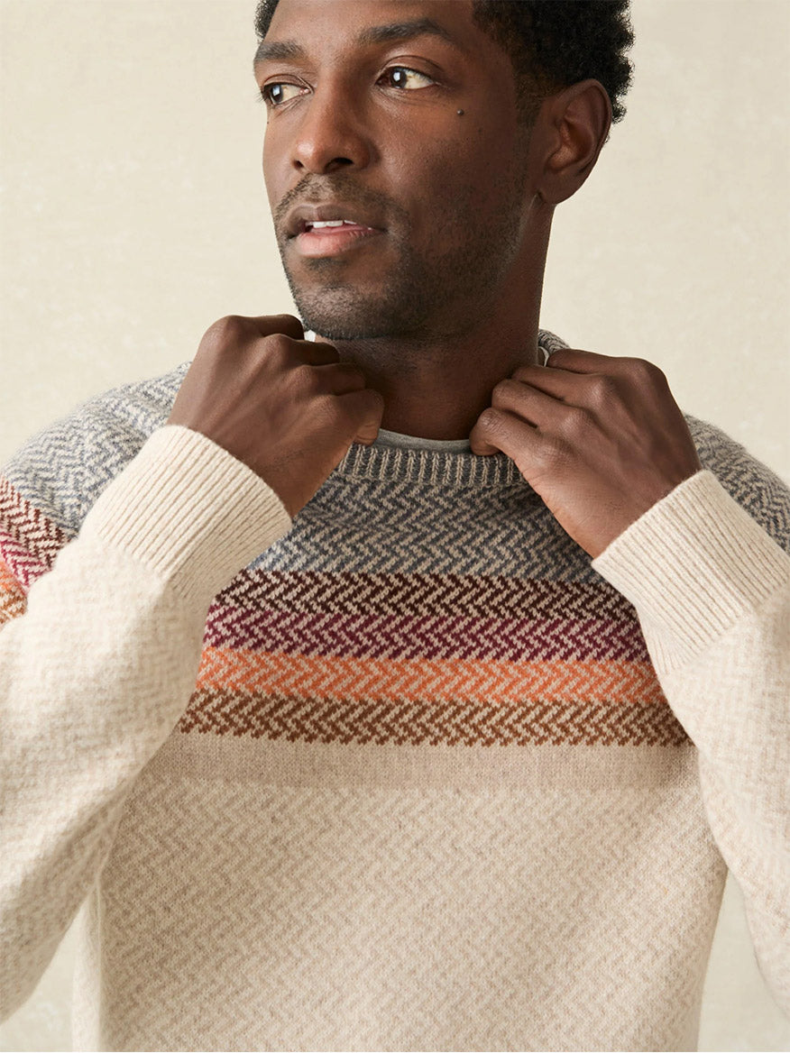 A person wearing the Faherty Brand Donegal Ombre Crew in Desert Sunset Ombre, a cream winter sweater made from a soft wool-blend and featuring a multi-colored striped pattern around the chest and shoulders.