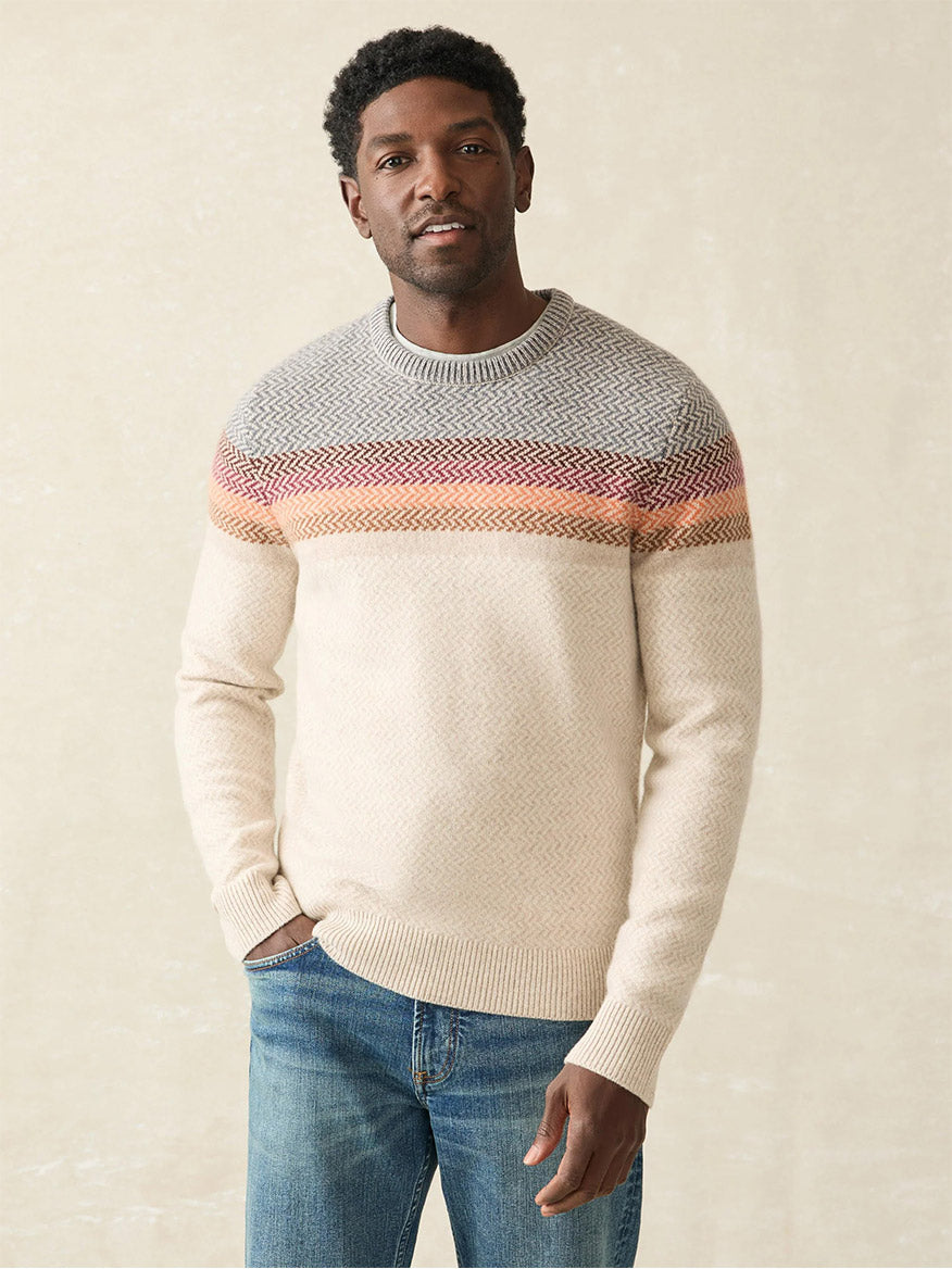 A person poses wearing the Faherty Brand Donegal Ombre Crew in Desert Sunset Ombre, showcasing colorful patterns on the upper part. It's paired with blue jeans against a neutral background.