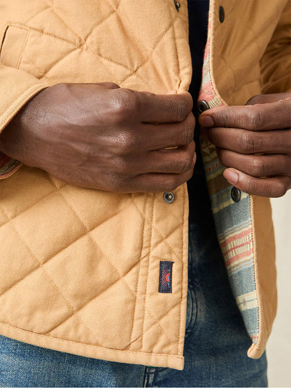 A person buttons a quilted tan jacket featuring a patterned Doug Good Feather design lining, paired with jeans, wearing the Faherty Brand Doug Good Feather Reversible Bondi Jacket in Dried Palm Eldorado Springs.