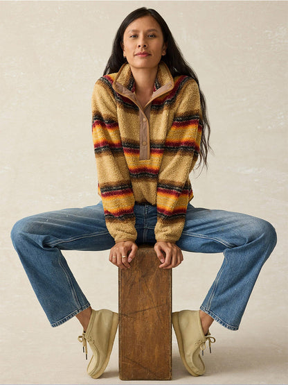 A person clad in a Faherty Brand Fireside Fleece Popover in Bonfire Stripe and jeans is seated on a wooden block against a plain background, ready for outdoor adventures.