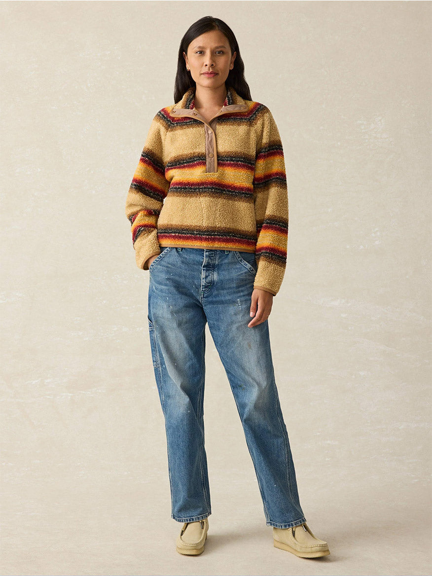 A person stands against a plain background, enveloped in the cozy warmth of the Faherty Brand Fireside Fleece Popover in Bonfire Stripe, paired with blue jeans and beige shoes.