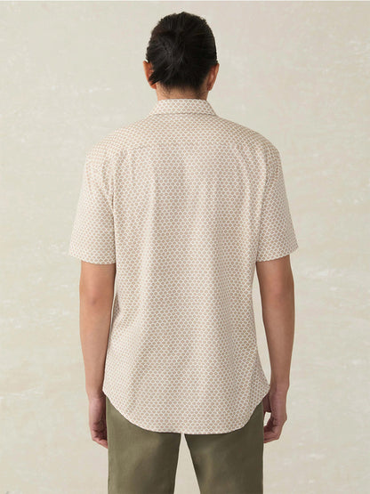 A person is shown from the back against a neutral background, wearing the Faherty Brand Short-Sleeve Hermosa Shirt in Island Sand Fishscale—a light-colored, printed Supima cotton shirt—paired with olive green pants.