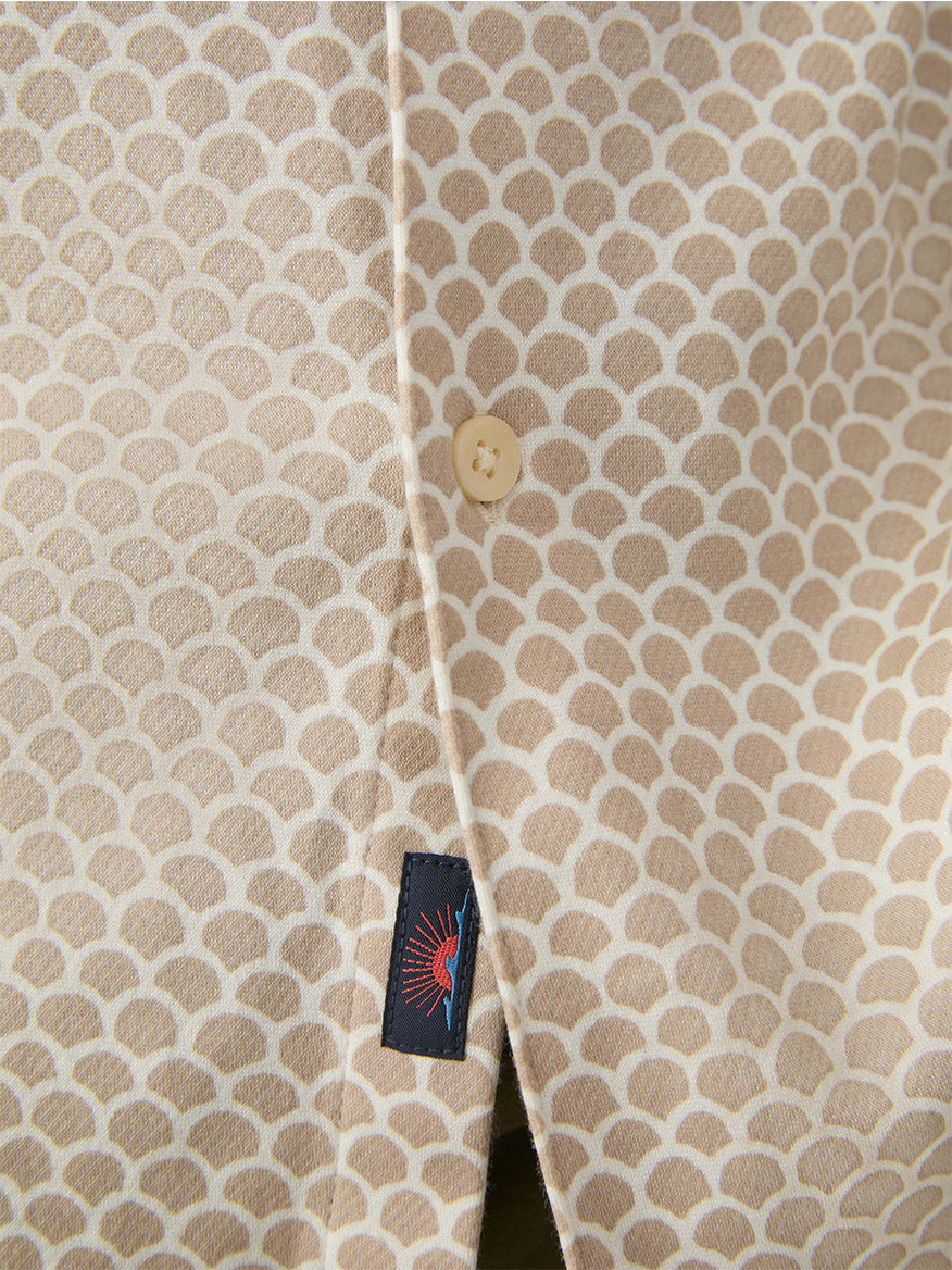 Close-up of the Faherty Brand Short-Sleeve Hermosa Shirt in Island Sand Fishscale—crafted from Supima cotton, featuring a scallop pattern, beige button, and black tag with a red and blue sunburst for subtle travel-inspired style.