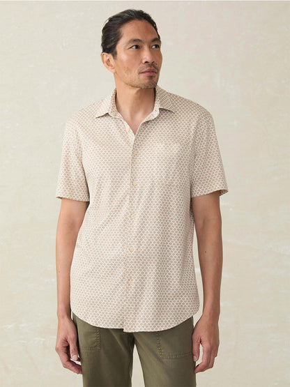 Man stands against a light background, wearing the Faherty Brand Short-Sleeve Hermosa Shirt in Island Sand Fishscale and olive green pants.