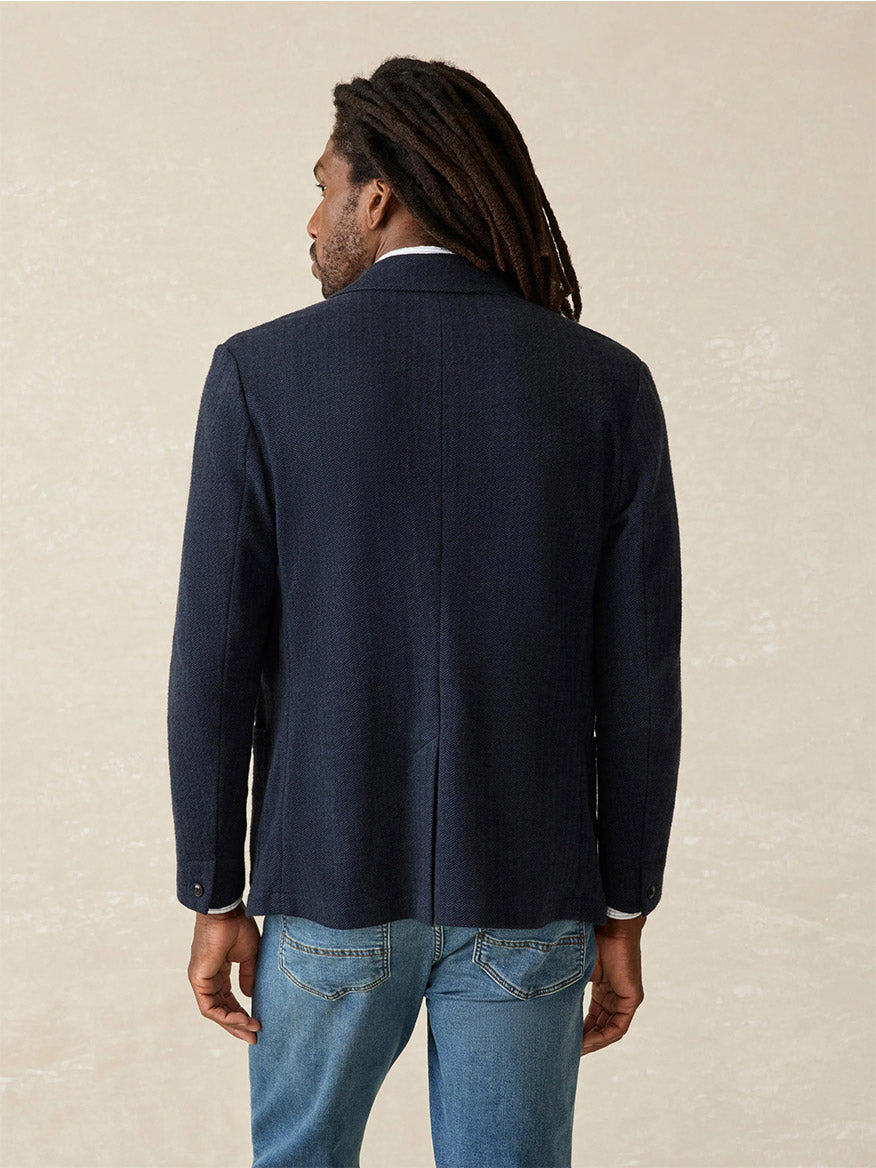 A man with long hair wearing the Faherty Brand Inlet Herringbone Blazer in Navy Twilight, paired with jeans, showcases a versatile layer of style as he faces away from the camera against a neutral background.