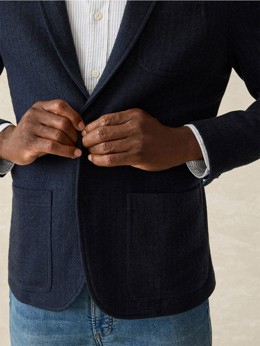 Dressed in a white shirt and blue jeans, a person fastens the Faherty Brand Inlet Herringbone Blazer in Navy Twilight, showcasing both style and versatility.